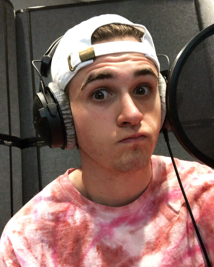 August Black wearing headphones, a white baseball cap, and a pink tie-dye shirt in the recording booth.