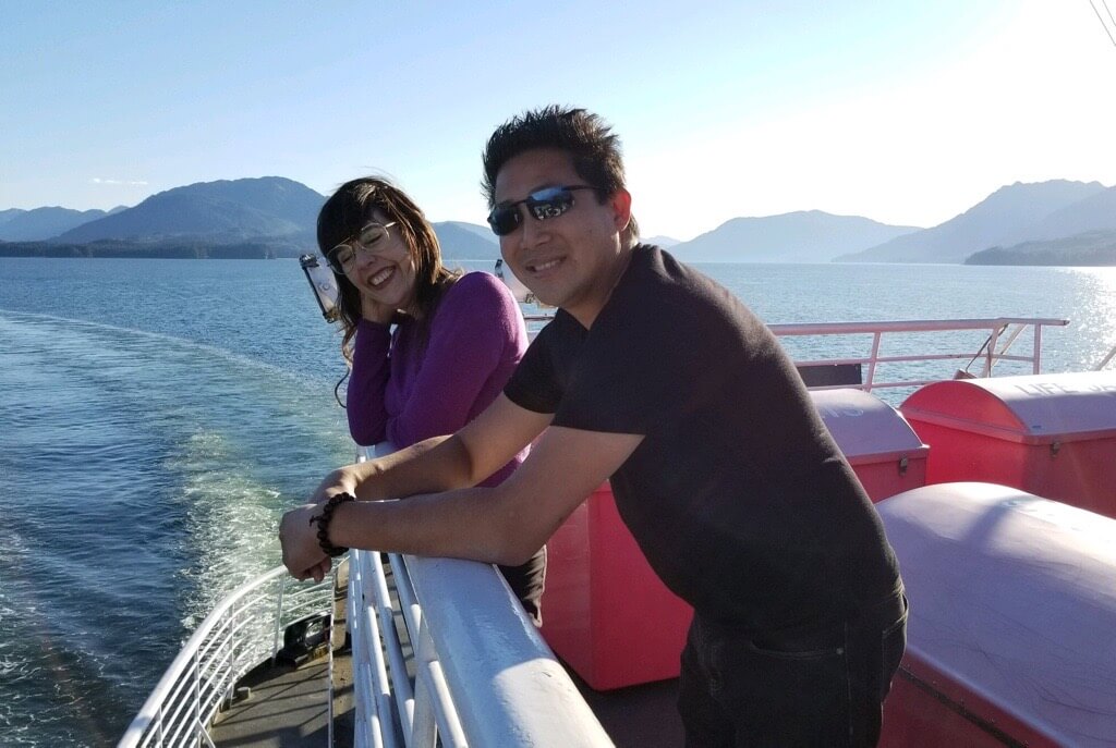 Elise Baldwin and Dennis Cheng smiling on a ferry boat with mountains behind them.